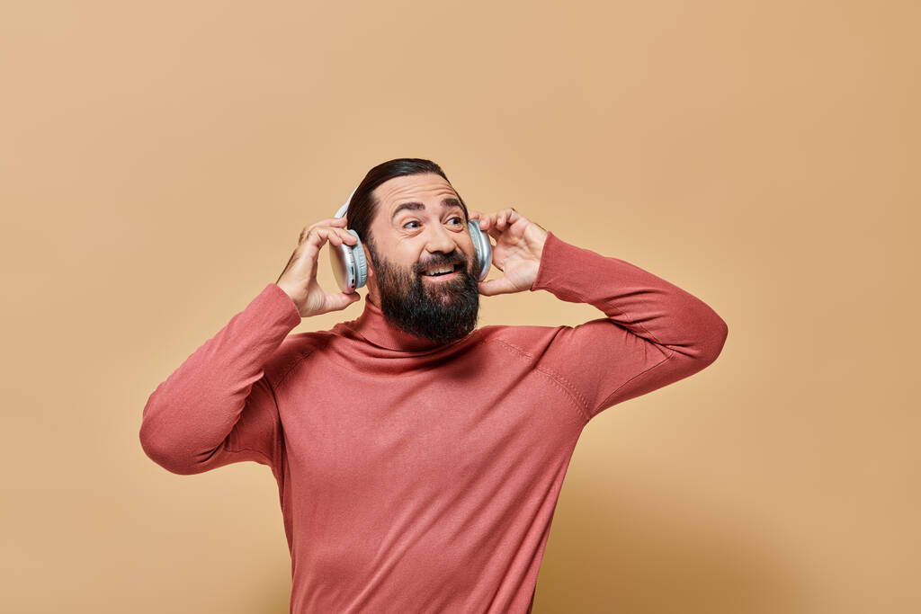 ευχαριστημένος γενειοφόρος άνδρας σε ζιβάγκο jumper ακούγοντας μουσική σε ασύρματα ακουστικά, μπεζ φόντο - Φωτογραφία, εικόνα