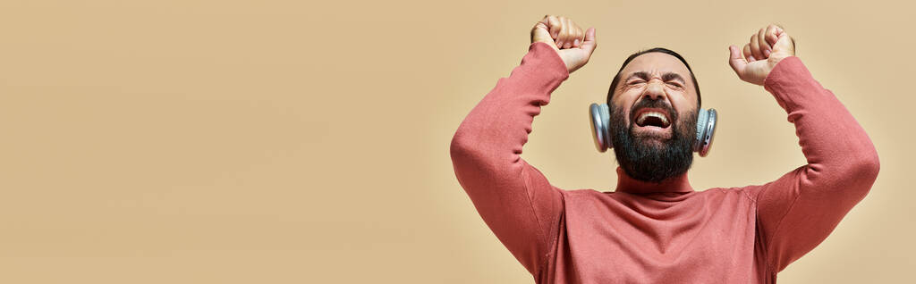 vzrušený vousatý muž v roláku jumper poslech hudby v bezdrátových sluchátkách, béžová, banner - Fotografie, Obrázek