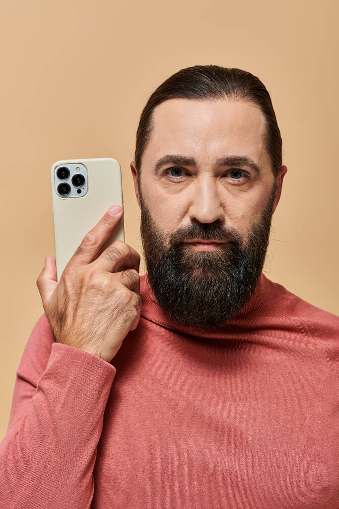 πορτρέτο του όμορφου γενειοφόρου άνδρα σε ζιβάγκο πουλόβερ κρατώντας smartphone σε μπεζ φόντο - Φωτογραφία, εικόνα