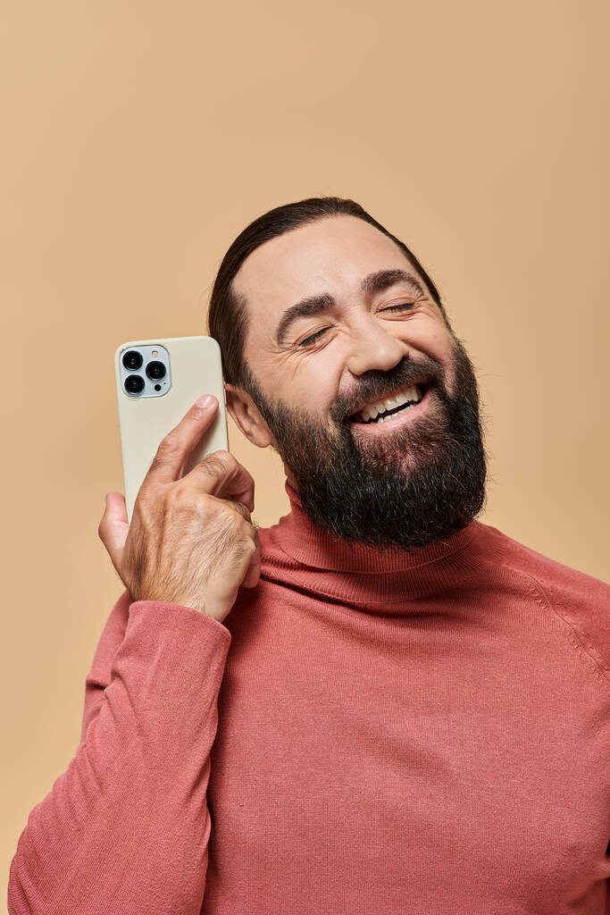 ベージュの背景にスマートフォンを保持するタートルネックジャンパーの幸せなひげ男の肖像画 - 写真・画像