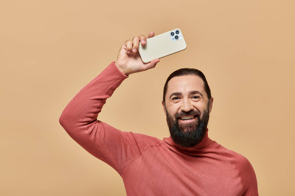 retrato de hombre barbudo feliz en jersey de cuello alto sosteniendo teléfono inteligente sobre fondo beige - Foto, imagen