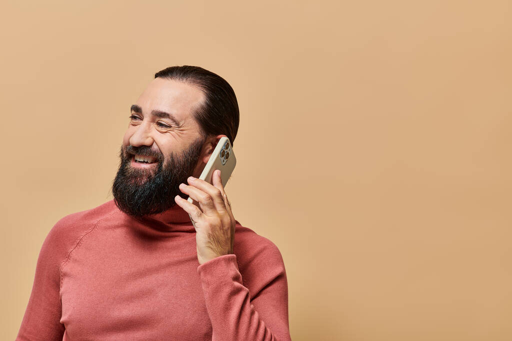 Porträt eines fröhlichen bärtigen Mannes im Rollkragenpullover, der auf einem Smartphone auf beigem Hintergrund spricht - Foto, Bild