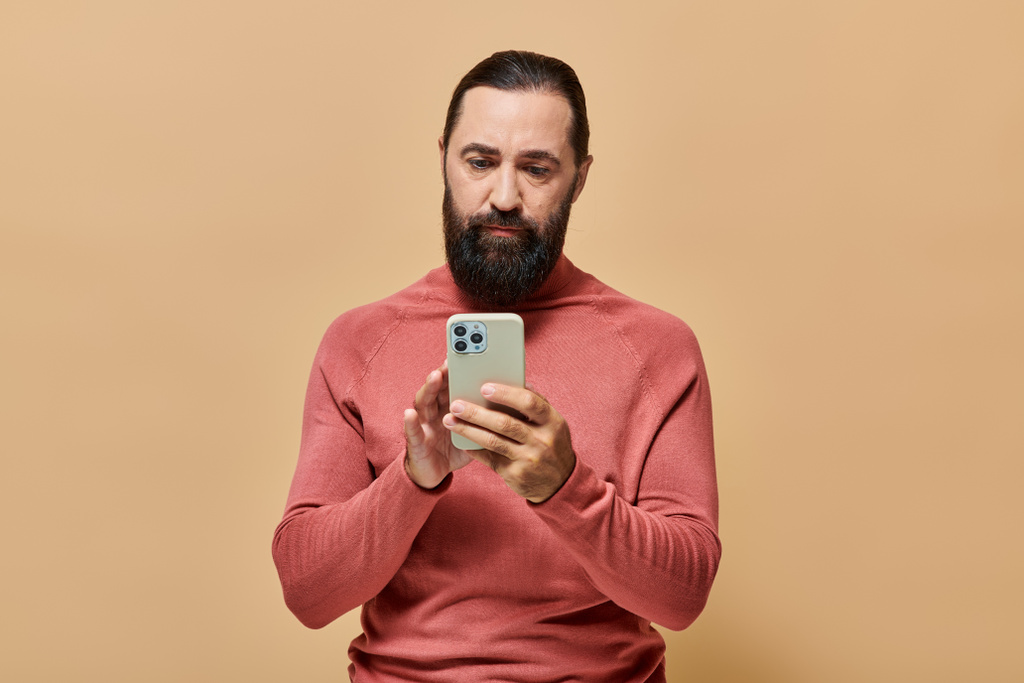 ritratto di bellissimo uomo barbuto in maglione dolcevita con smartphone su sfondo beige - Foto, immagini