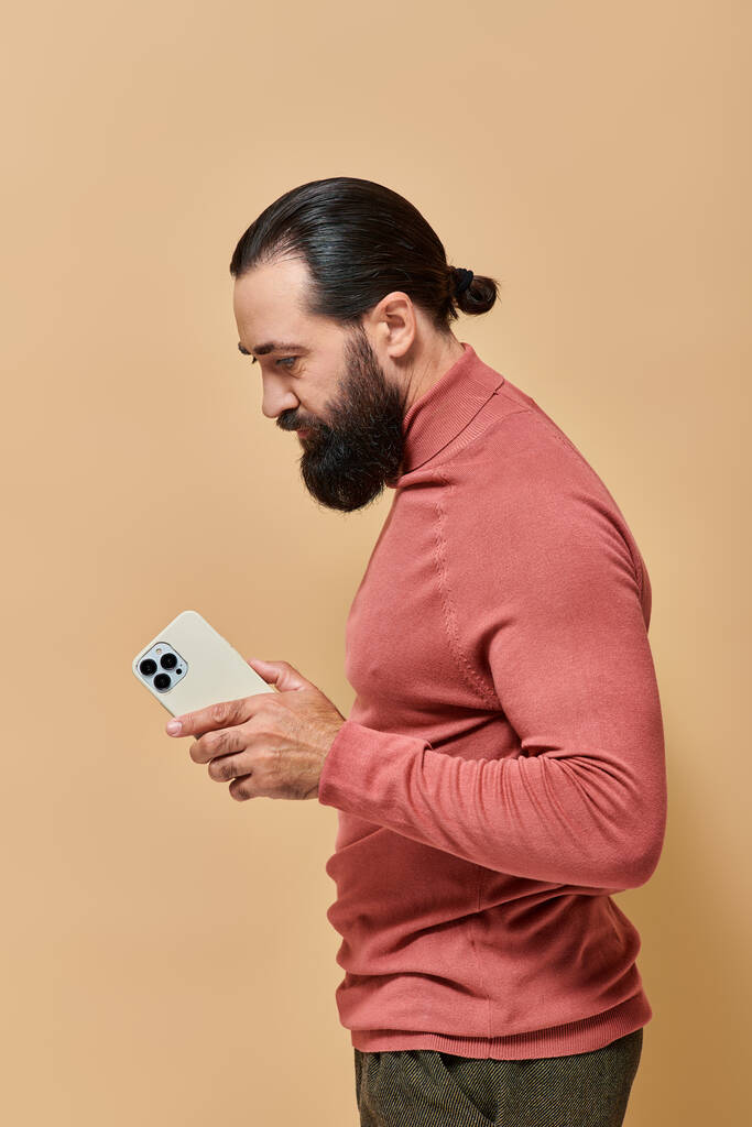 portrét vousatý dobře vypadající muž v roláku jumper držení smartphone na béžové pozadí - Fotografie, Obrázek