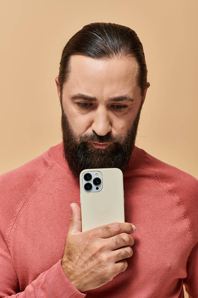 портрет хорошого виглядає бородатого чоловіка в стрибку з водоростями, фотографуючи на смартфоні, бежевий фон - Фото, зображення