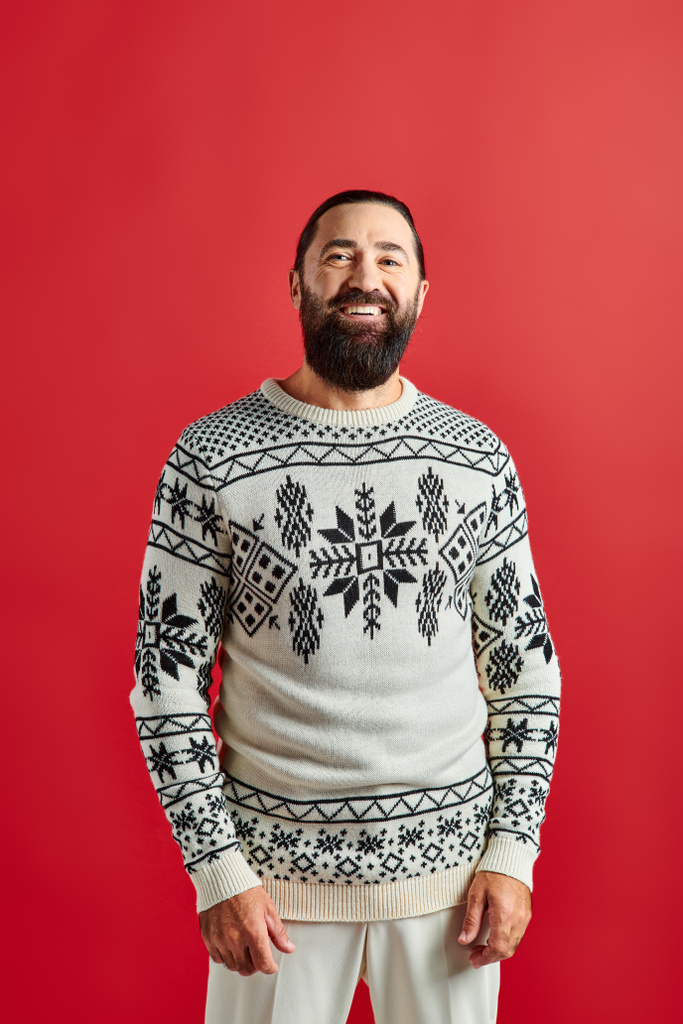 szczęśliwy brodaty mężczyzna w zimowym swetrze z ozdobą uśmiechnięty na czerwonym tle, Wesołych Świąt - Zdjęcie, obraz