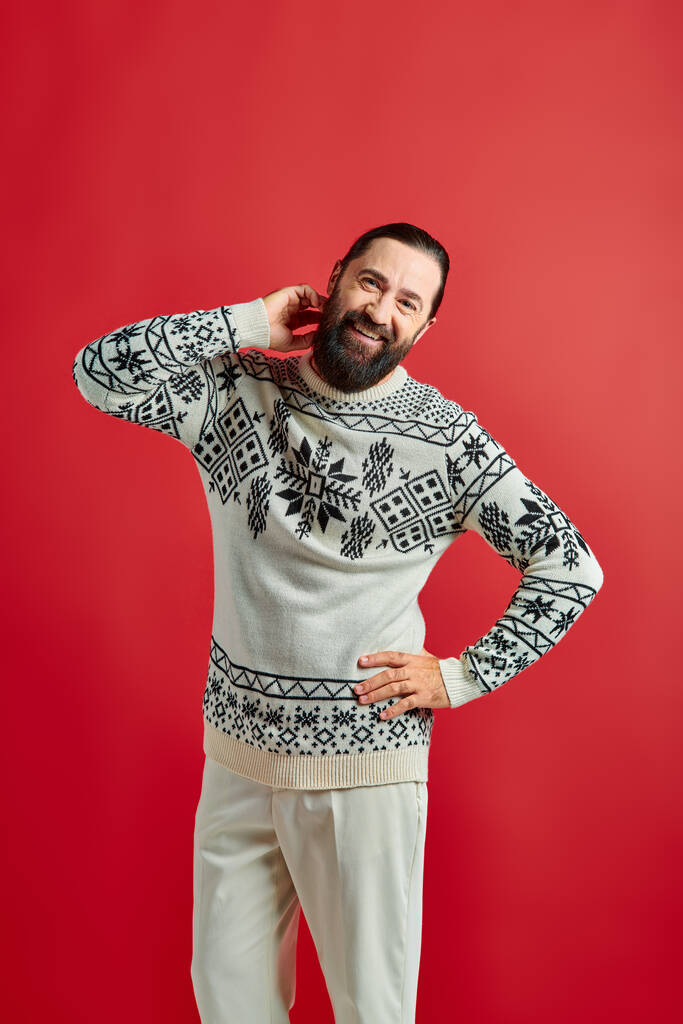 heureux homme barbu en pull d'hiver posant avec la main sur la hanche sur fond rouge, Joyeux Noël - Photo, image
