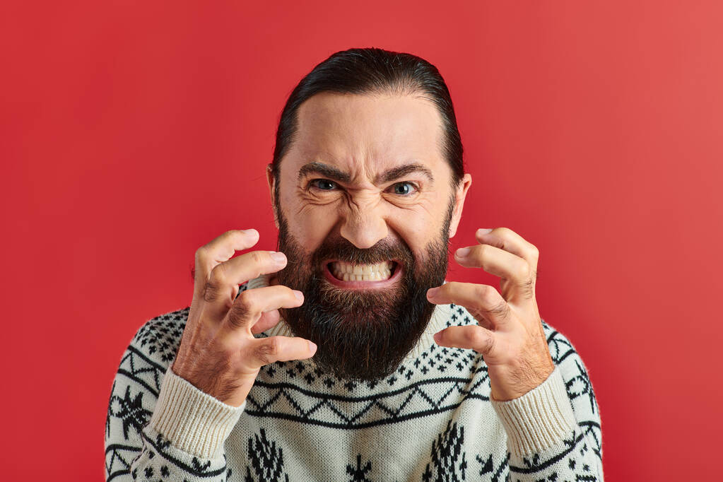angry bearded man in winter sweater with ornament grinning and gesturing on red background - Photo, Image