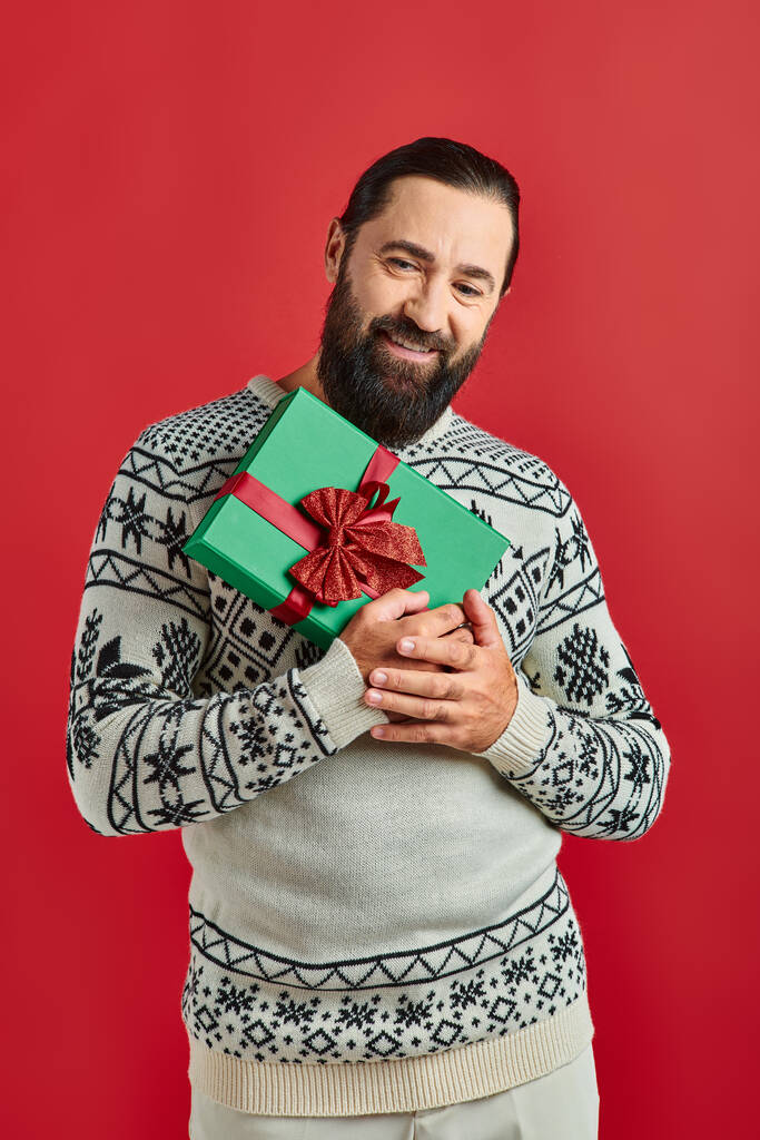 glücklich bärtiger Mann im Winterpullover mit Ornament hält Weihnachtsgeschenk auf rotem Hintergrund - Foto, Bild