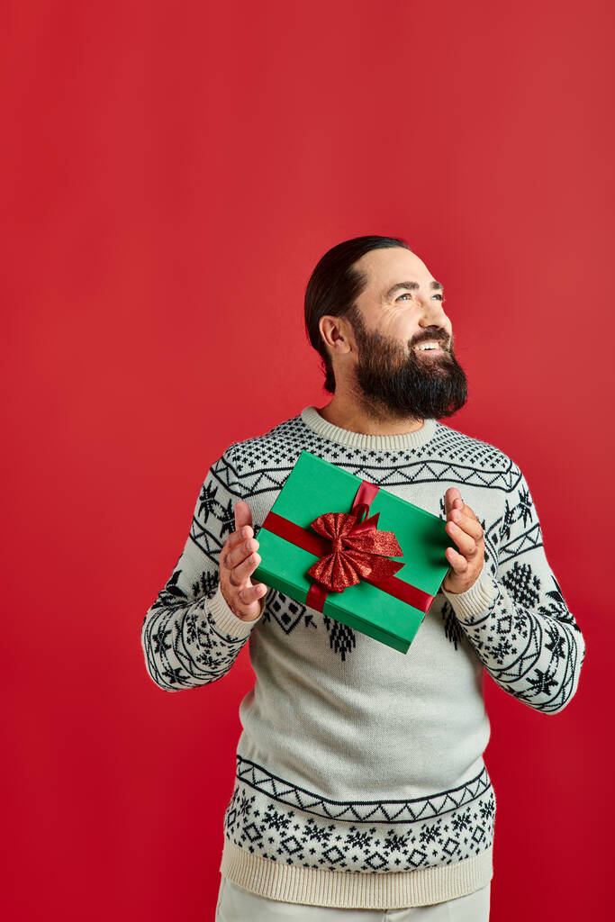 щасливий бородатий чоловік в зимовому светрі з прикрасою, що тримає різдвяний подарунок на червоному тлі - Фото, зображення