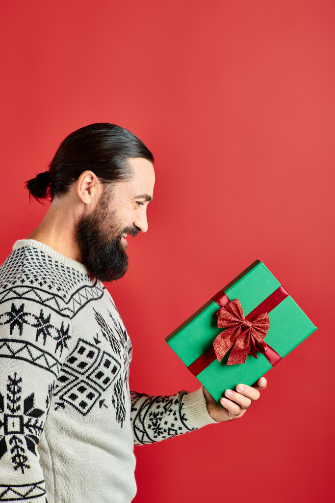 vista lateral, hombre barbudo feliz en suéter de invierno con adorno celebración regalo de Navidad en rojo - Foto, imagen