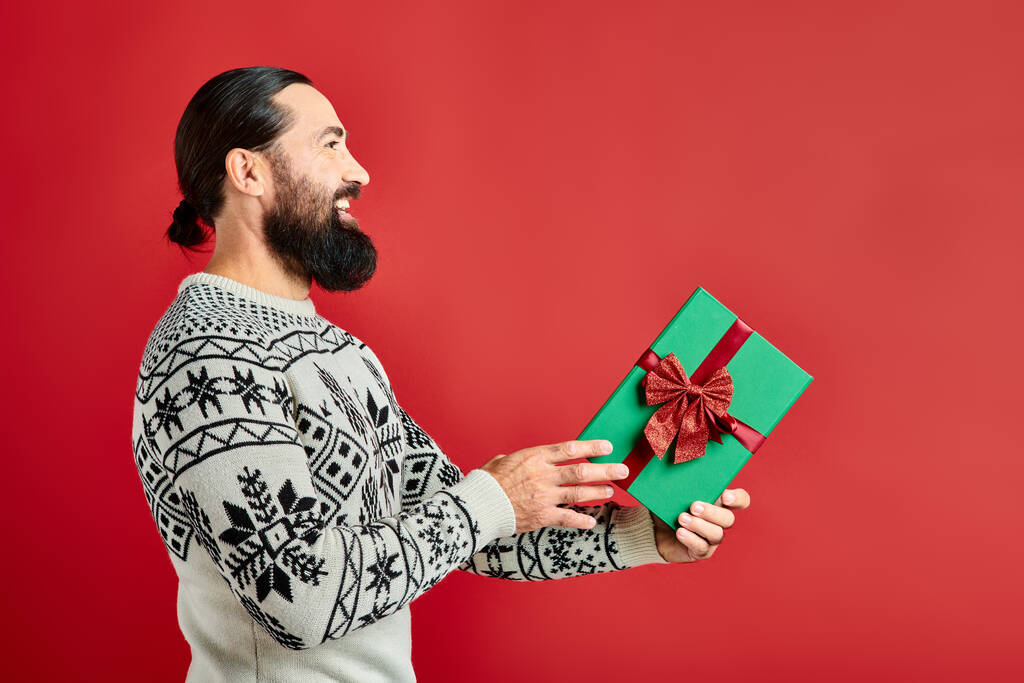 side view, pleased bearded man in winter sweater with ornament holding Christmas present on red - Photo, Image