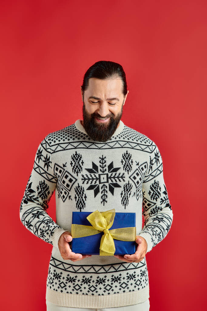 zadowolony brodaty mężczyzna w zimowym swetrze z ozdobą gospodarstwa prezent świąteczny na czerwonym tle - Zdjęcie, obraz