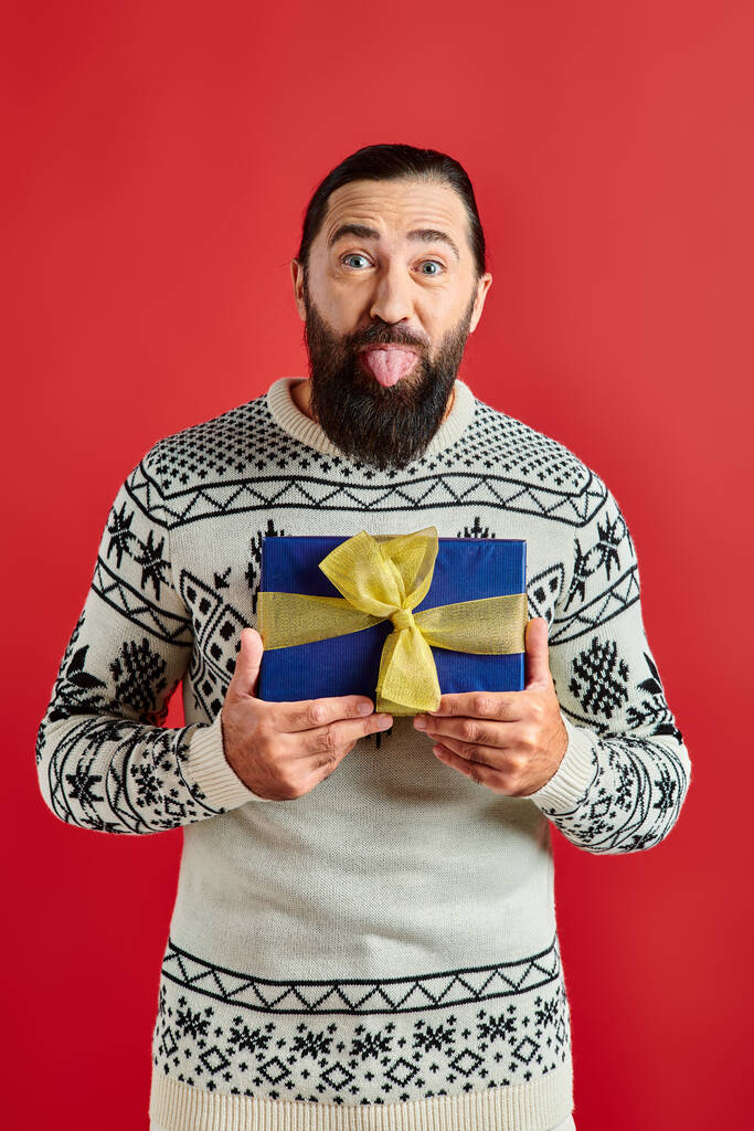 смешной бородатый мужчина в зимнем свитере с орнаментом держа рождественский подарок и приклеивая язык на красный - Фото, изображение