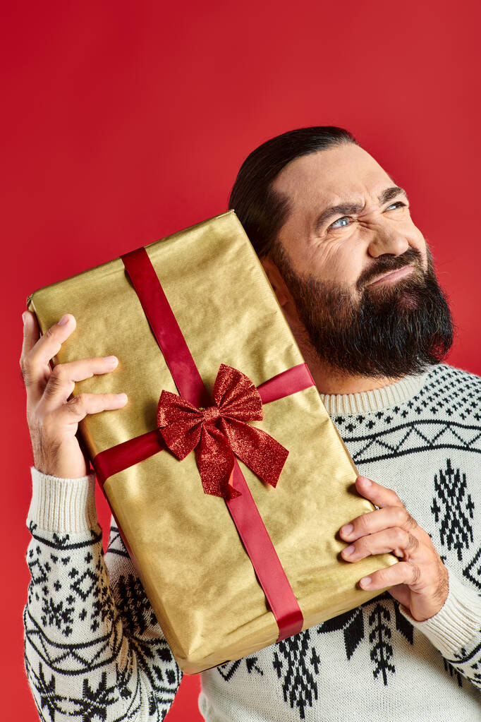 Kış süveteri giymiş sakallı, hoşnutsuz bir adam. Kırmızı arka planda Noel hediyesi tutuyor. - Fotoğraf, Görsel