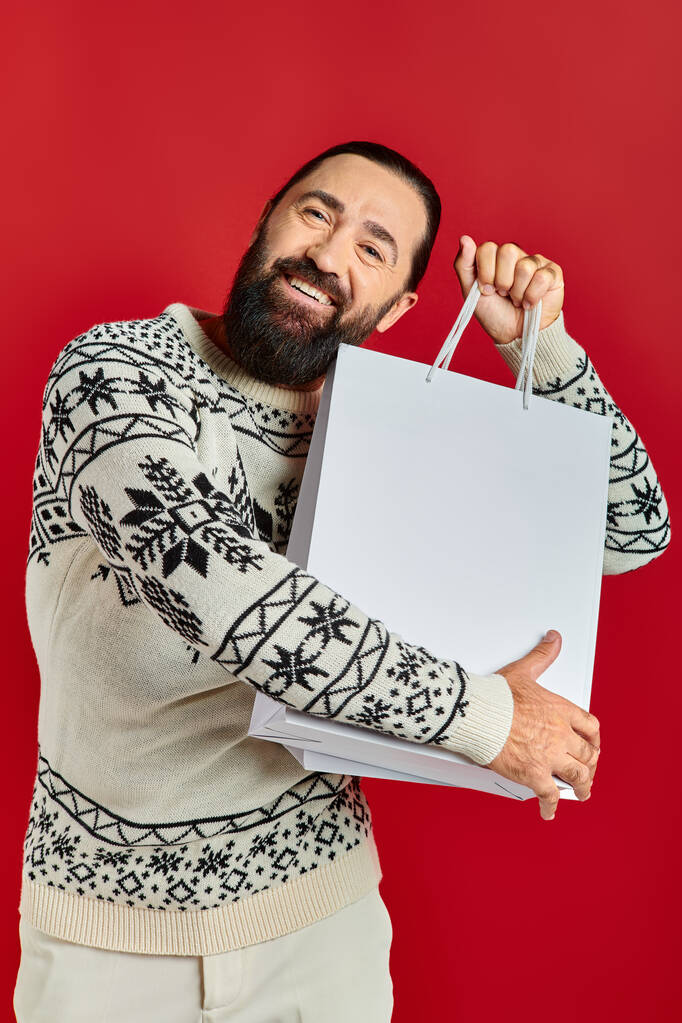 homem feliz barbudo em camisola de Natal segurando sacos de compras em pano de fundo vermelho, vendas de férias - Foto, Imagem