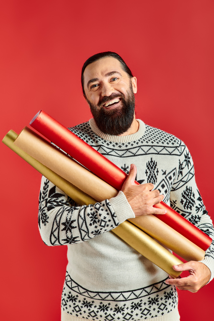 χαρούμενος γενειοφόρος άνδρας σε χριστουγεννιάτικο πουλόβερ κρατώντας χαρτί περιτυλίγματος σε κόκκινο φόντο, πωλήσεις διακοπών - Φωτογραφία, εικόνα