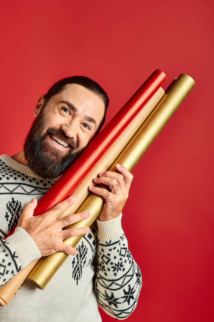 hombre barbudo feliz en suéter de invierno sosteniendo papel de embalaje sobre fondo rojo, Feliz Navidad - Foto, imagen