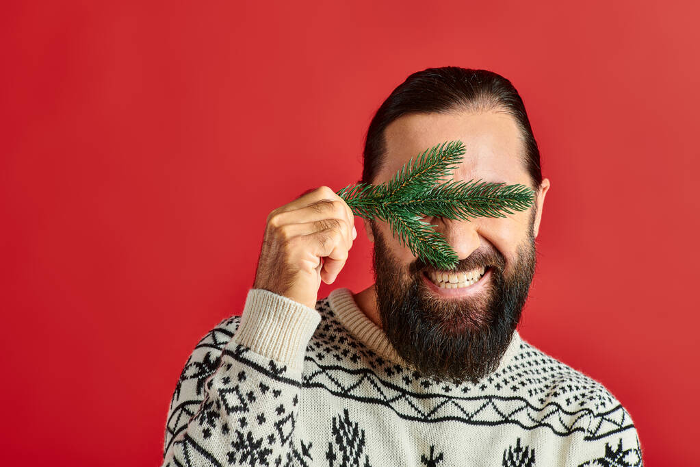 joyful man in sweater covering eyes with branch of pine tree on red backdrop, Merry Christmas - Photo, Image
