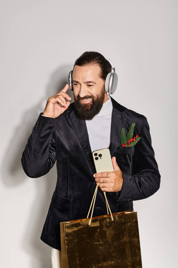 šťastný muž poslouchá hudbu v bezdrátových sluchátkách, drží smartphone a dárkové tašky na šedém pozadí - Fotografie, Obrázek
