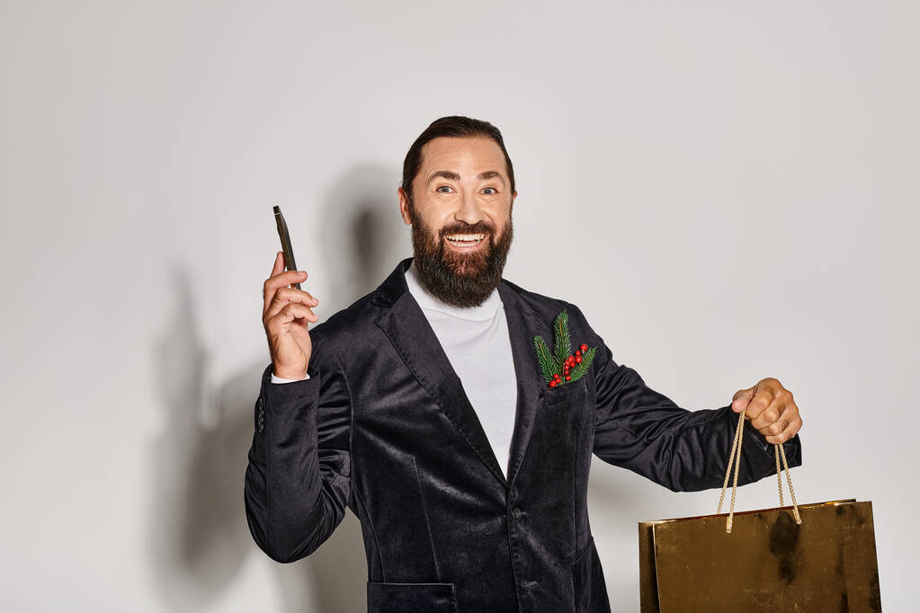 счастливый бородатый человек в костюме держа смартфон и рождественский подарочный пакет на сером фоне, покупки - Фото, изображение