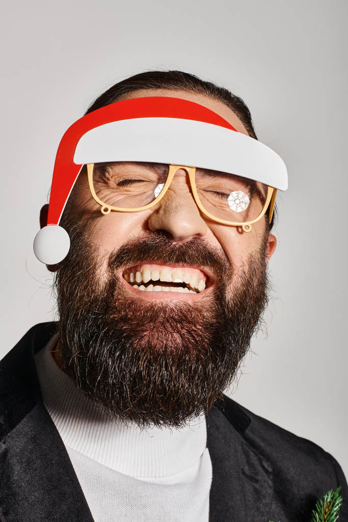 радісний бородатий чоловік у святкових окулярах з капелюхом Санти позує в костюмі і посміхається на сірому фоні - Фото, зображення