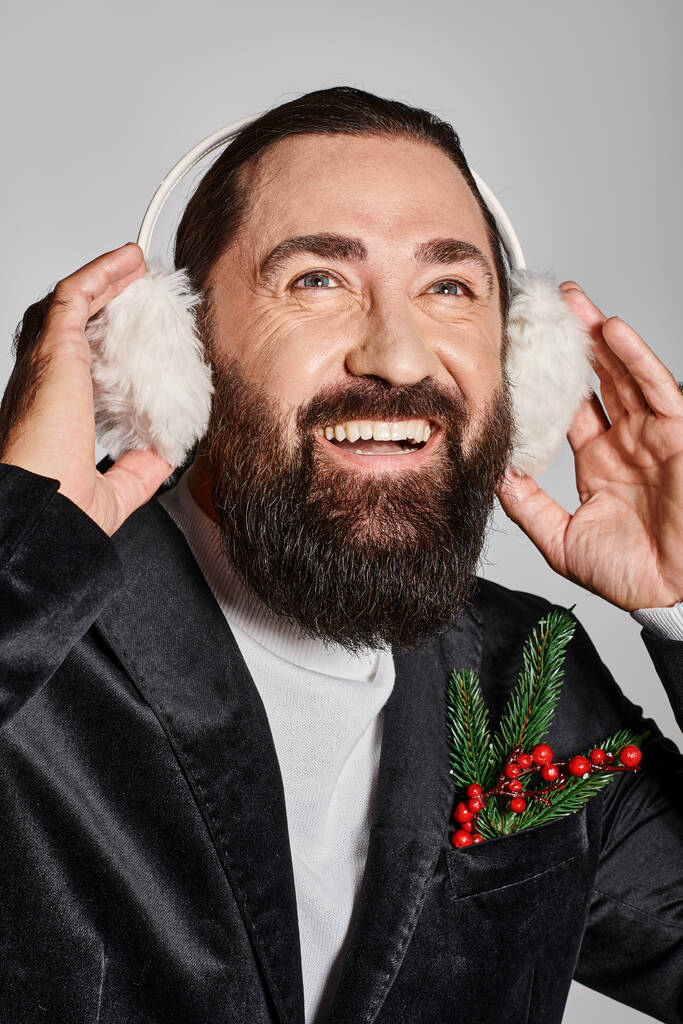 счастливый бородатый мужчина в костюме с еловой веткой и красными ягодами в наушниках на сером - Фото, изображение