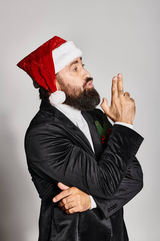 portret brodatego przystojniaka w formalnym stroju i czapce Mikołaja pozującego na szarym tle, Boże Narodzenie - Zdjęcie, obraz