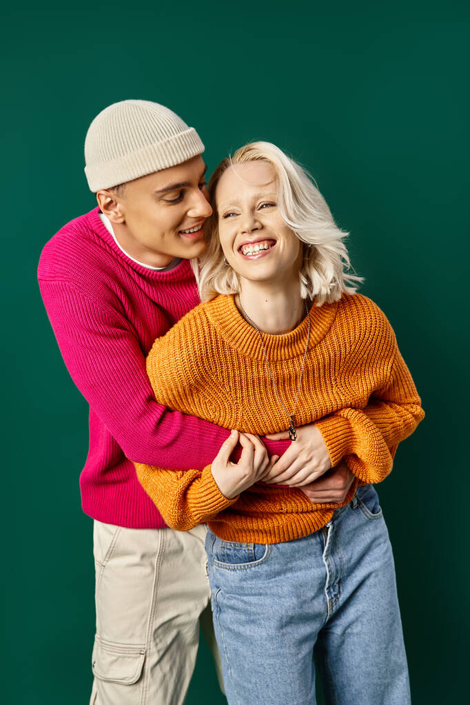 Paar in Winterkleidung amüsiert sich, Mann in Mütze umarmt glückliche Freundin vor türkisfarbener Kulisse - Foto, Bild