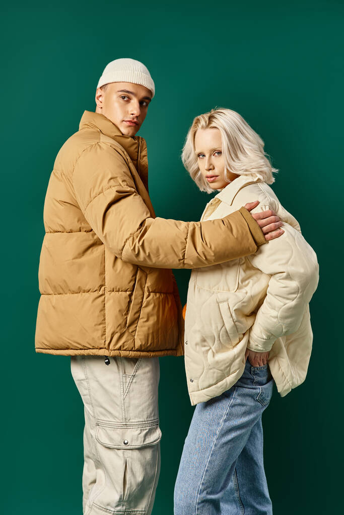 стильная пара в куртках фугу позируют вместе на бирюзовом фоне, молодой человек и женщина - Фото, изображение
