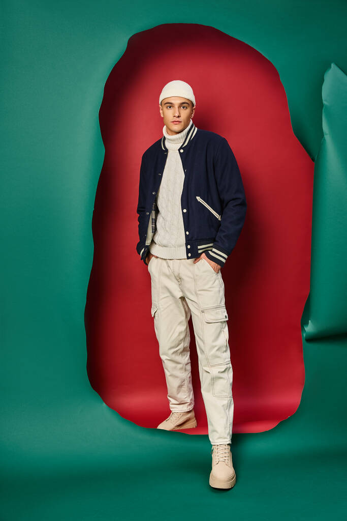 stijlvolle jongeman in beanie, witte trui en bomberjack op rood gescheurd met turquoise achtergrond - Foto, afbeelding