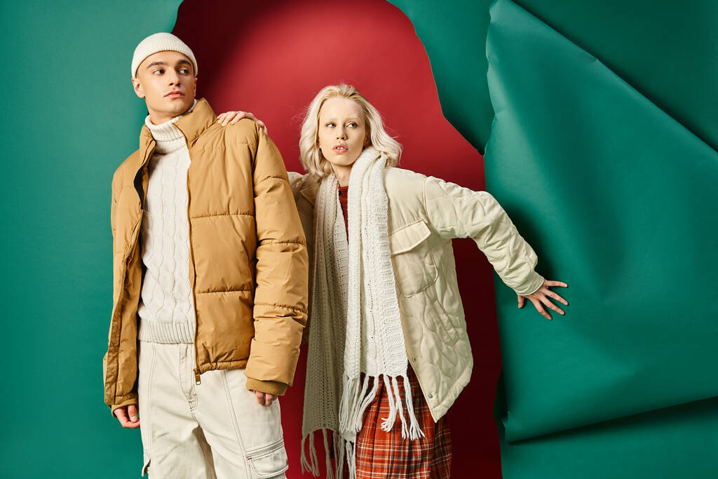 стильный молодой человек и женщина в зимних куртках позируют вместе на красном фоне с бирюзовым - Фото, изображение