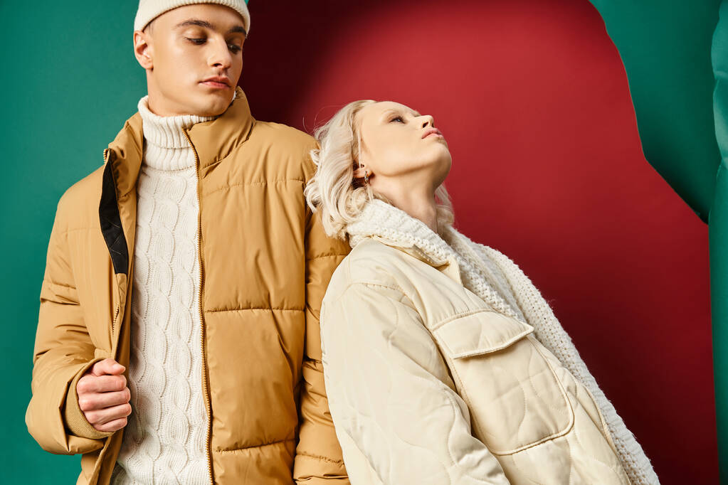 blonde woman in winter jacket leaning on handsome man in beanie on red with turquoise backdrop - Photo, Image
