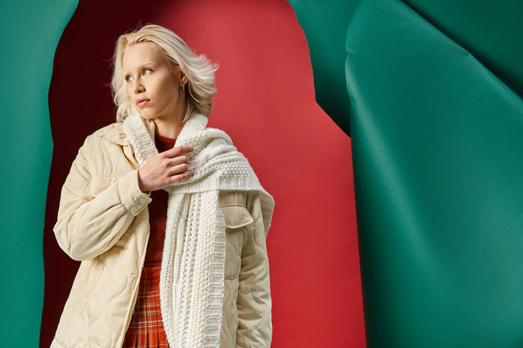 冬のジャケットの若い金髪の女性はターコイズの背景で赤に編まれたスカーフを調整 - 写真・画像