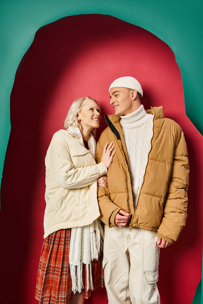 冬のジャケットで幸せな女性は,引き裂かれたターコイズと赤い背景の近くで陽気なボーイフレンドを抱擁 - 写真・画像