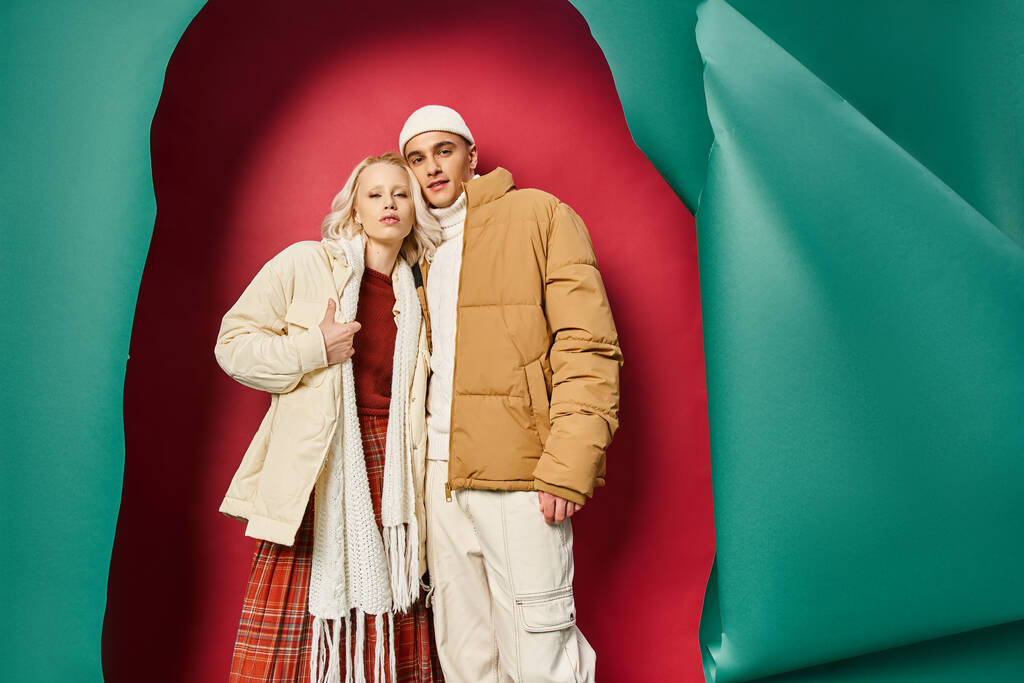 стильная молодая пара в теплой зимней верхней одежде позируют вместе возле разорванной бирюзы и красный фон - Фото, изображение