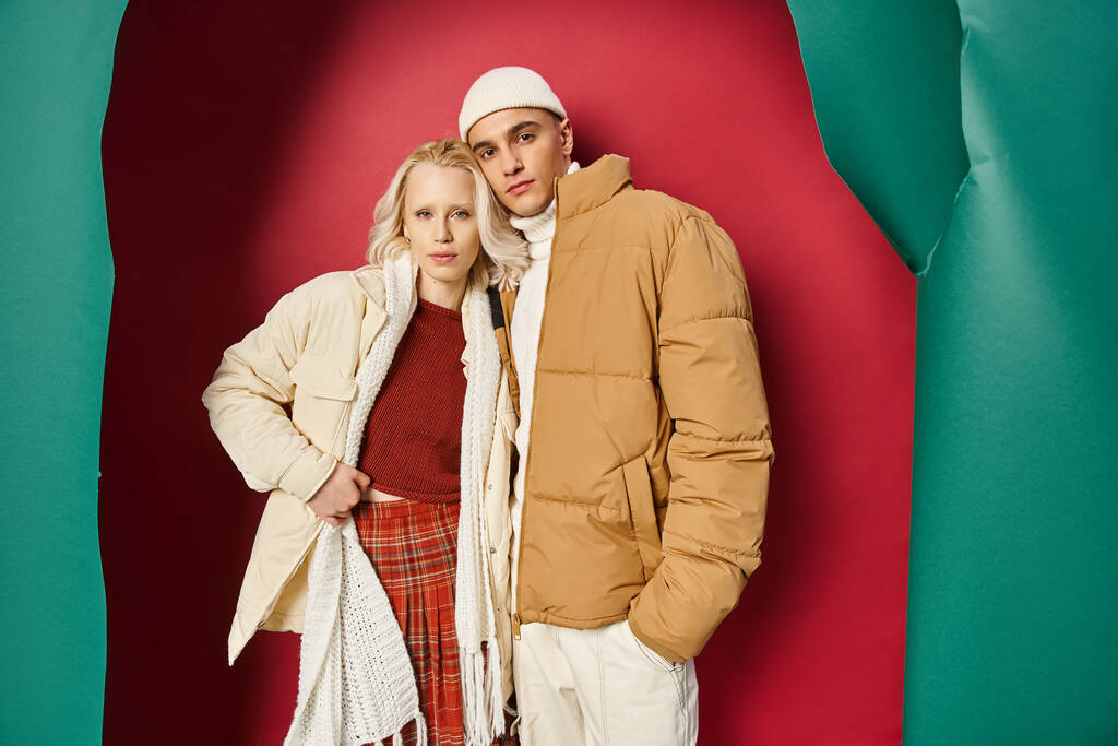 atraktivní žena a muž v zimě svrchní oblečení pózovat společně v blízkosti roztržené tyrkysové a červené pozadí - Fotografie, Obrázek
