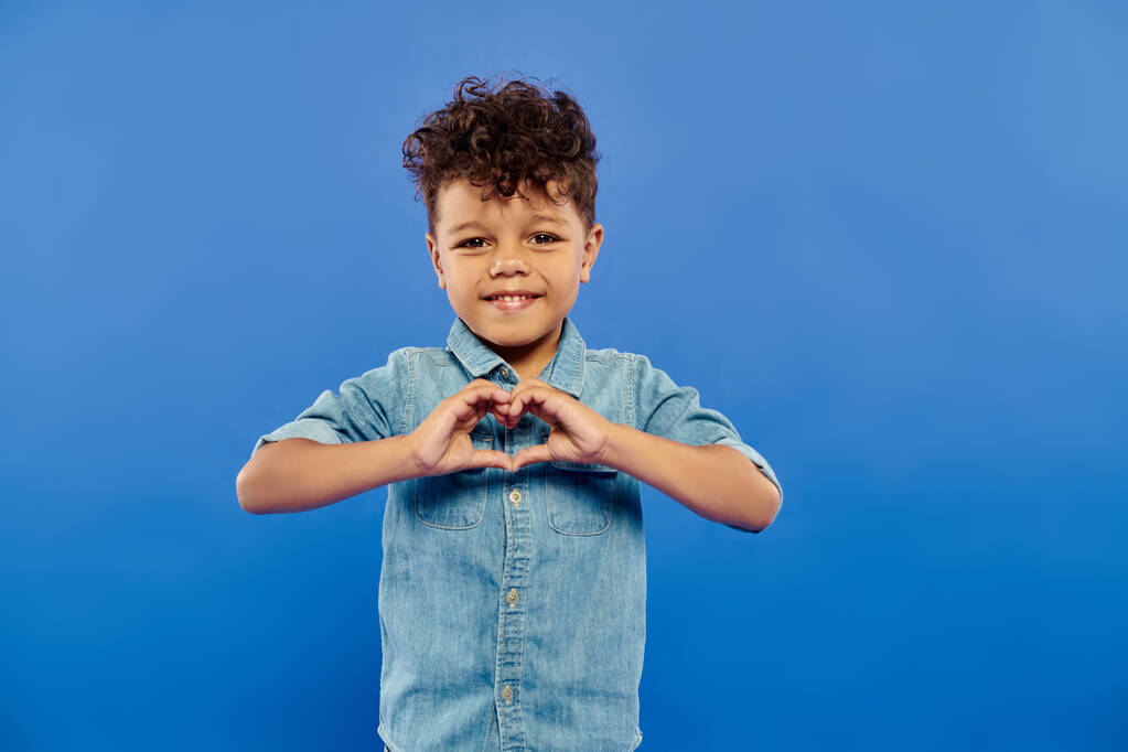 happy african american preschooler boy in stylish denim outfit showing heart with hands on blue - Photo, Image