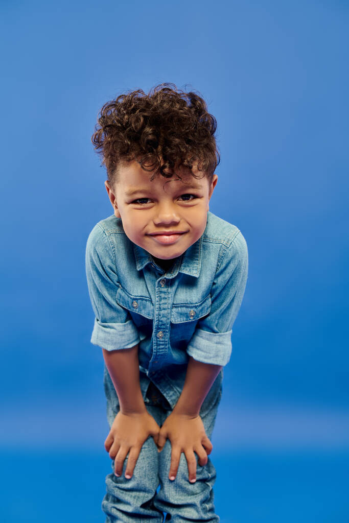 pequeno menino pré-escolar afro-americano feliz em roupa de ganga elegante posando no fundo azul - Foto, Imagem