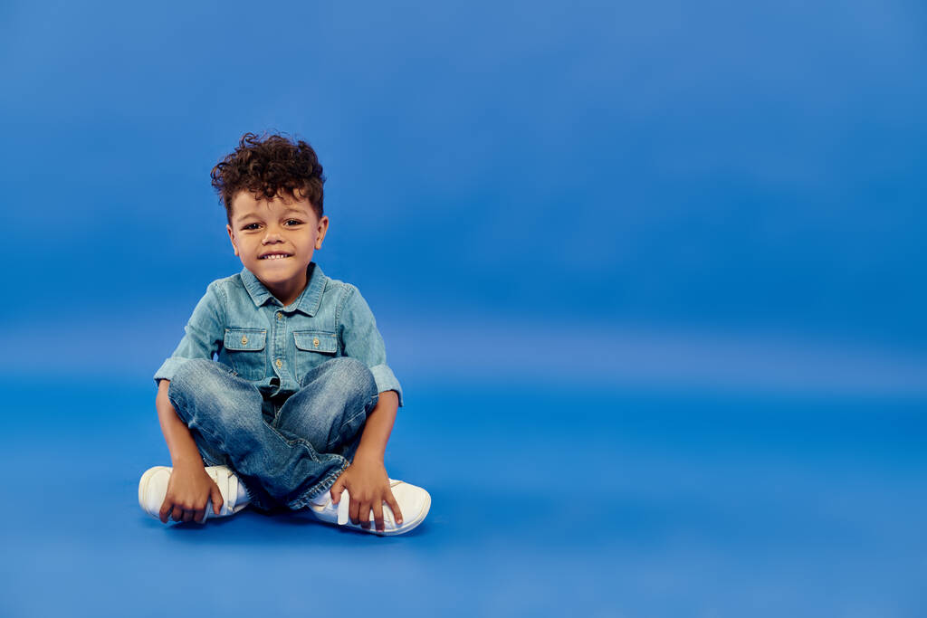 счастливый кудрявый африканский дошкольник в стильном джинсовом наряде сидит на синем фоне - Фото, изображение
