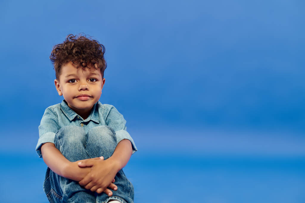 позитивный африканский американский дошкольник в стильной джинсовой одежде сидит на синем фоне - Фото, изображение
