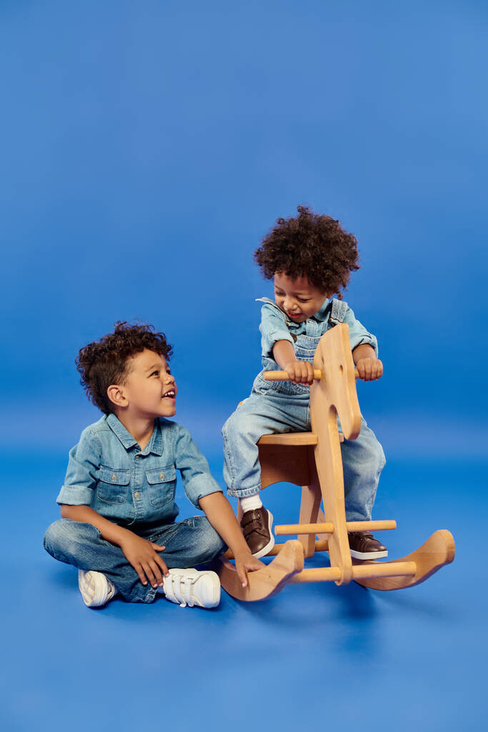 cute african american toddler boy in stylish denim clothes sitting on rocking horse near brother - Photo, Image