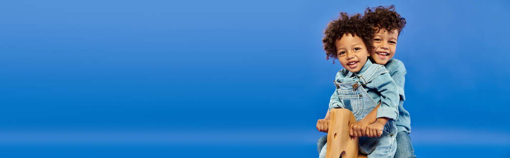 fröhliche afrikanisch-amerikanische Kinder in stylischer Jeanskleidung auf Schaukelpferd auf blauem Banner sitzend - Foto, Bild