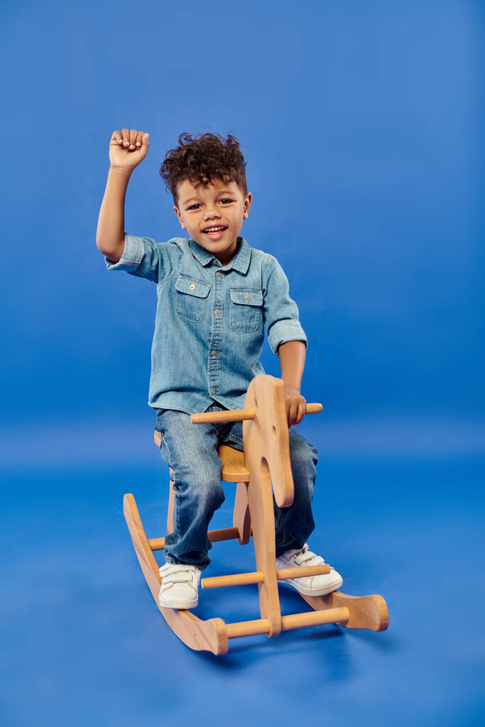 cute african american preschooler boy in stylish denim clothes sitting on rocking horse on blue - Photo, Image