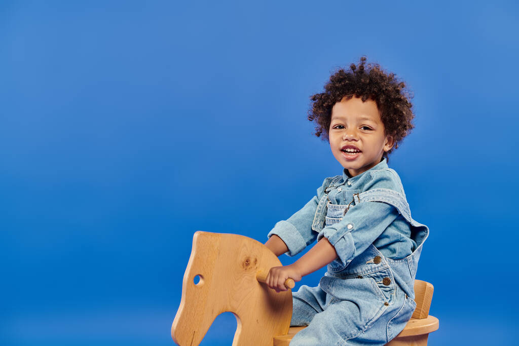 gelukkig afrikaans amerikaans peuter jongen in denim kleding zitten op schommelstoel op blauw achtergrond - Foto, afbeelding
