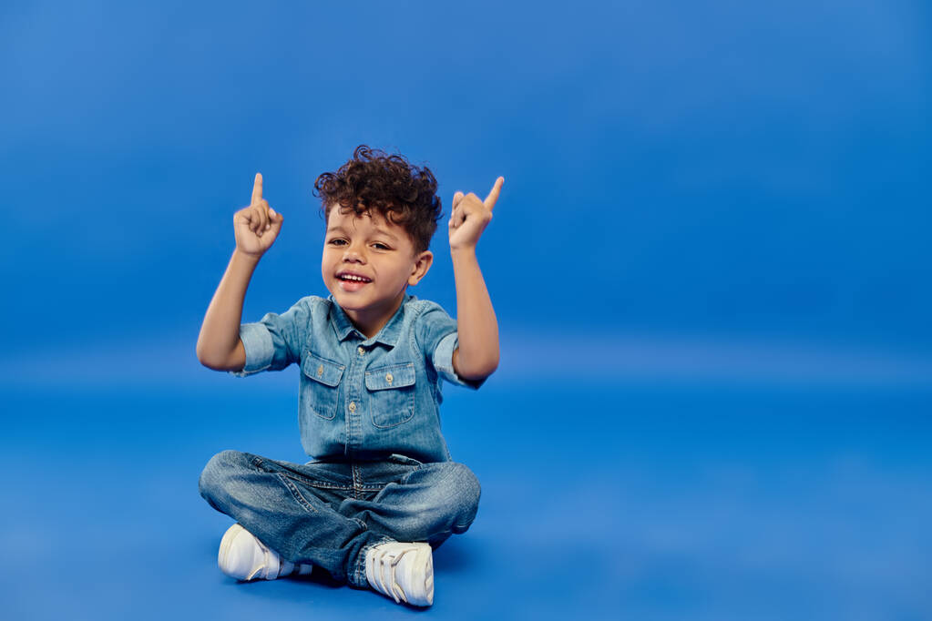 alegre y rizado afroamericano preescolar chico en ropa de mezclilla sentado apuntando hacia arriba en azul - Foto, Imagen