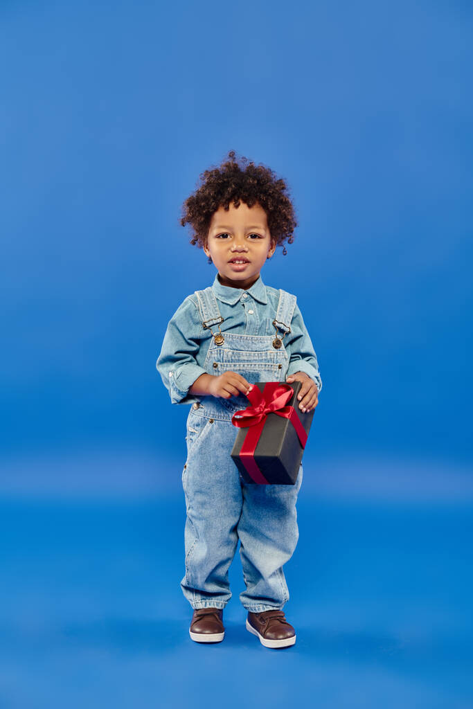 青い背景にクリスマスプレゼントと一緒に立っているデニム服の幸せなアフリカ系アメリカ人の少年 - 写真・画像