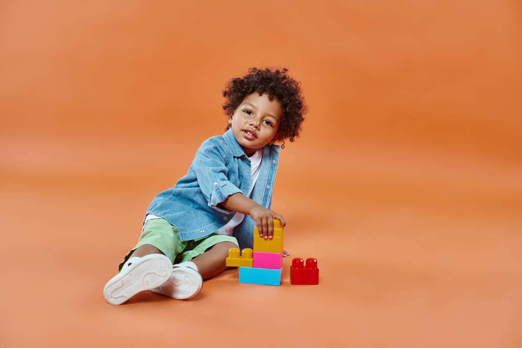かわいいアフリカ系アメリカ人の幼い少年 デニムシャツに座ってオレンジ色のブロックを構築 - 写真・画像