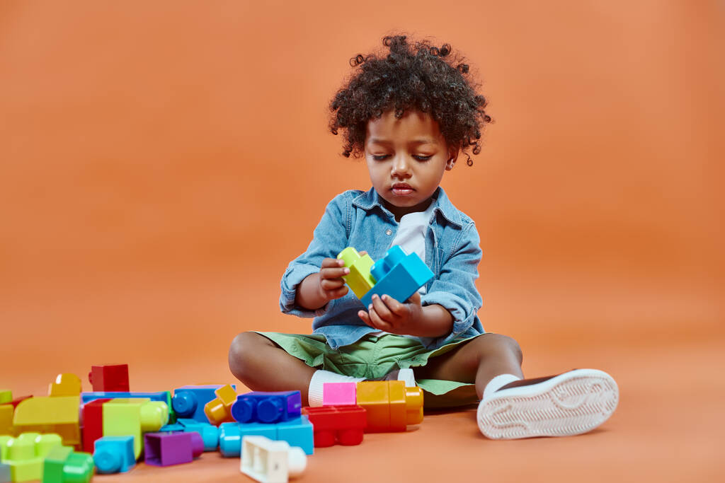 imádnivaló afro-amerikai csecsemő fiú alkalmi öltözékben ül és játszik építőkockák narancs - Fotó, kép