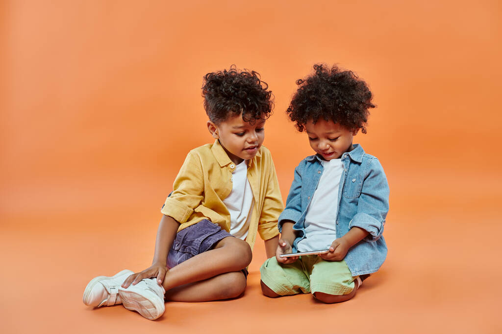 fröhliches afrikanisch-amerikanisches Kind in lässiger Kleidung beim Anblick des auf dem Smartphone spielenden Kleinkindes - Foto, Bild